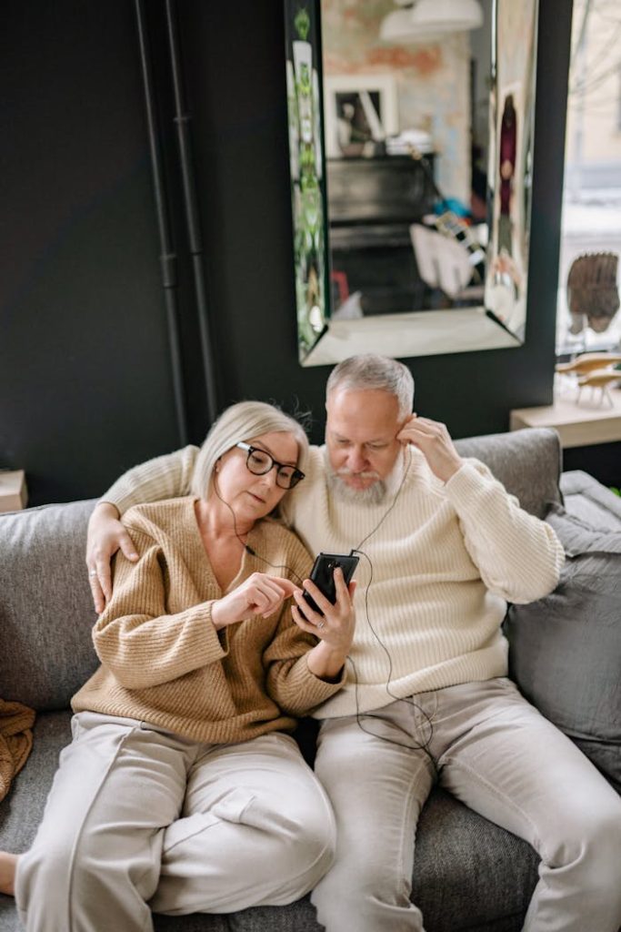 A happy senior couple listening to music together on a smartphone while sitting on a cozy sofa at home.